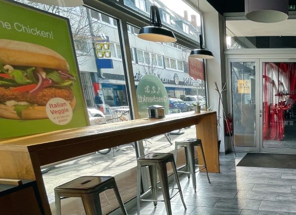 Innenbereich eines burgerme Restaurants mit Sitzgelegenheiten und italienischem Veggie-Burger auf Werbeplakat am Fenster.