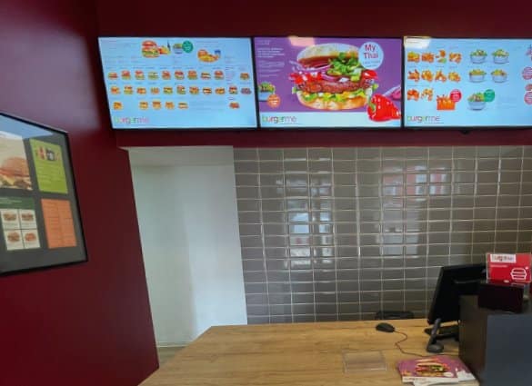 Digitale Menütafeln bei burgerme zeigen verschiedene Burger-Optionen über dem Bestellbereich in einem modernen Restaurant.