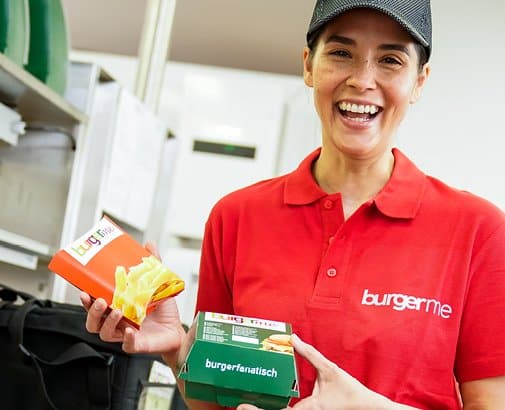 Mitarbeiterin von burgerme in rotem Polohemd hält Pommes und Burger-Verpackungen lächelnd im burgerme-Store