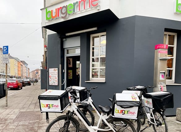 Burgerme Filiale mit Lieferfahrrädern im Vordergrund vor dem Geschäftseingang in einer belebten städtischen Straße.
