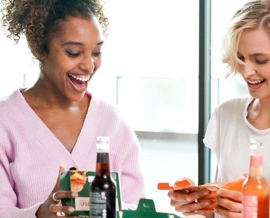 Zwei Frauen genießen ein leckeres Essen von burgerme mit Softdrinks in gemütlicher Atmosphäre.