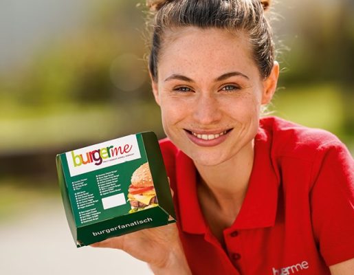 Mitarbeiterin in rotem Shirt hält eine burgerme-Verpackung in die Kamera und lächelt.